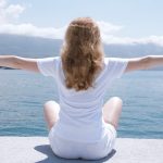Woman sitting with open arms by the water, embracing nature and serenity.