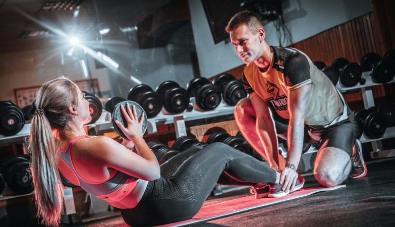A personal trainer assists a woman doing sit-ups with a medicine ball in a gym setting.